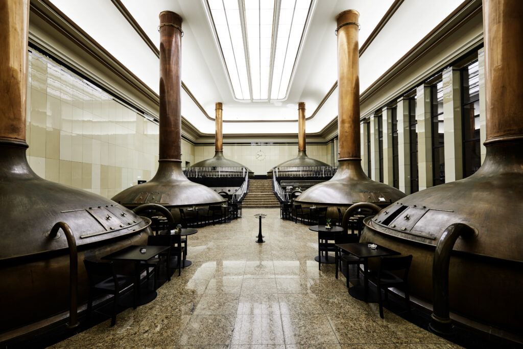 Erkennbar sind mehrere Meter große Kupferpfannen, deren Rohre bis unter die hohe Decke reichen. In der Mitte des Bildes führt eine Treppe auf eine Empore mit Geländer.