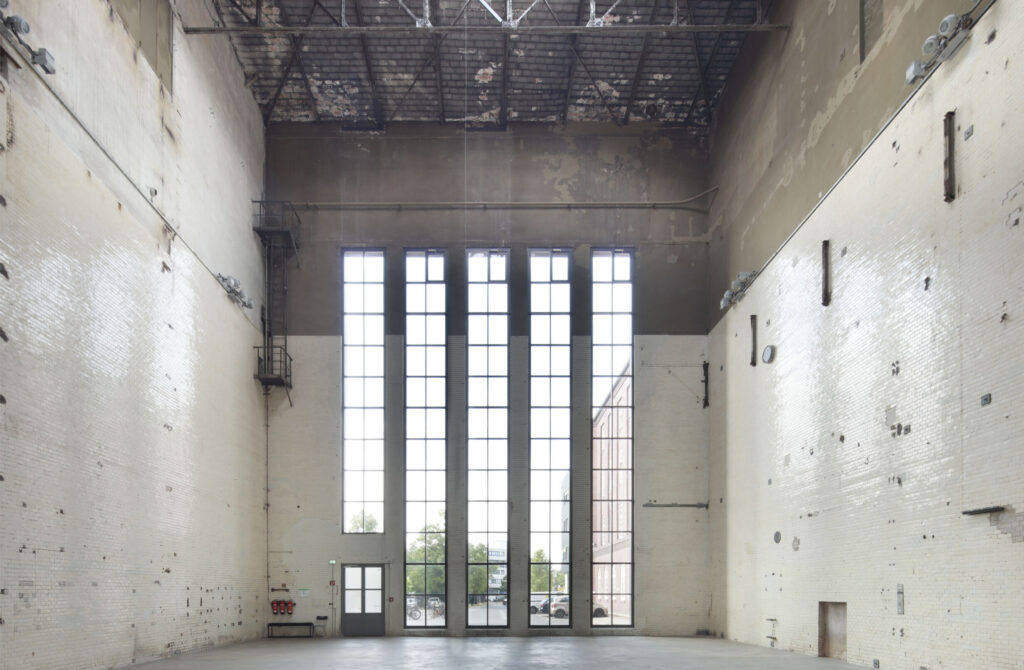 Blick in eine hohe Industrieshalle mit sehr großen Fenstern und glatten Wänden.