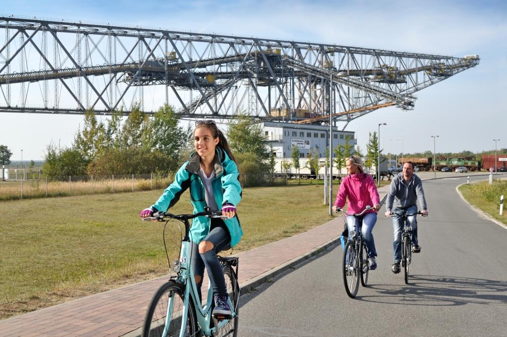 Drei Fahrradfahrer:innen auf der Straße vor der Förderbrücke.