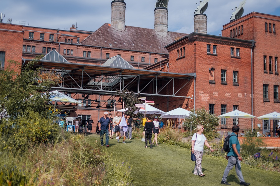 Menschen besuchen die Malzfabrik im Sommer. Sonnenschirme spenden Schatten.