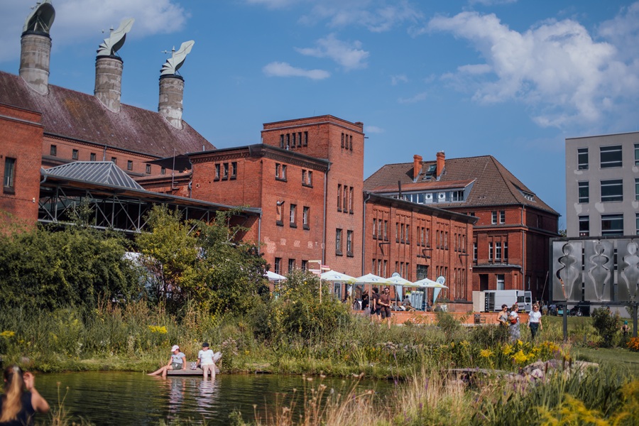 Am Ufer des Sees an der malzfabrik sitzen Kinder. Im Hintergrund ist ein großer Backsteinbau.
