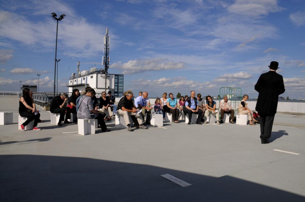 Theateraufführung im öffentlichen Raum. Das Publikum sitzt auf Hockern auf einem Parkdeck.