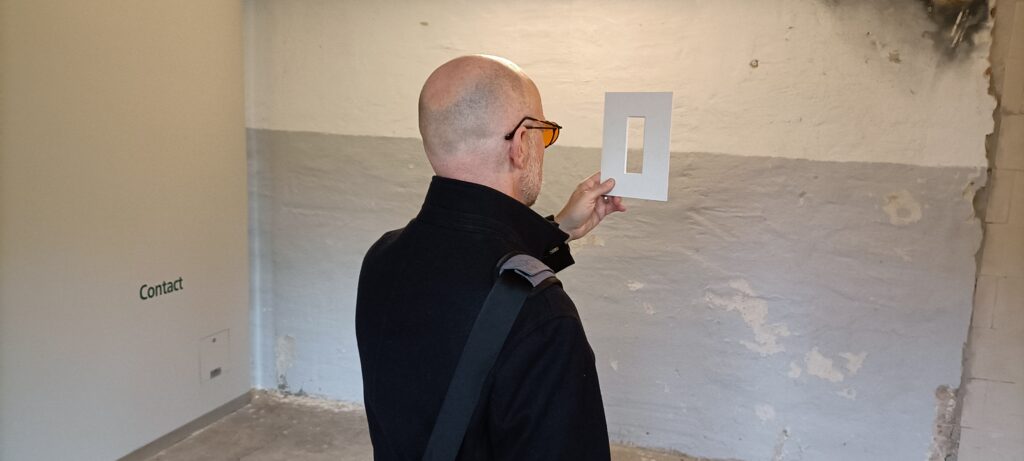 Ein Mann hält ein Passepartout vor sein Gesicht und blickt durch die Öffnung auf eine Wand im NS-Zwangsarbeiterlager.