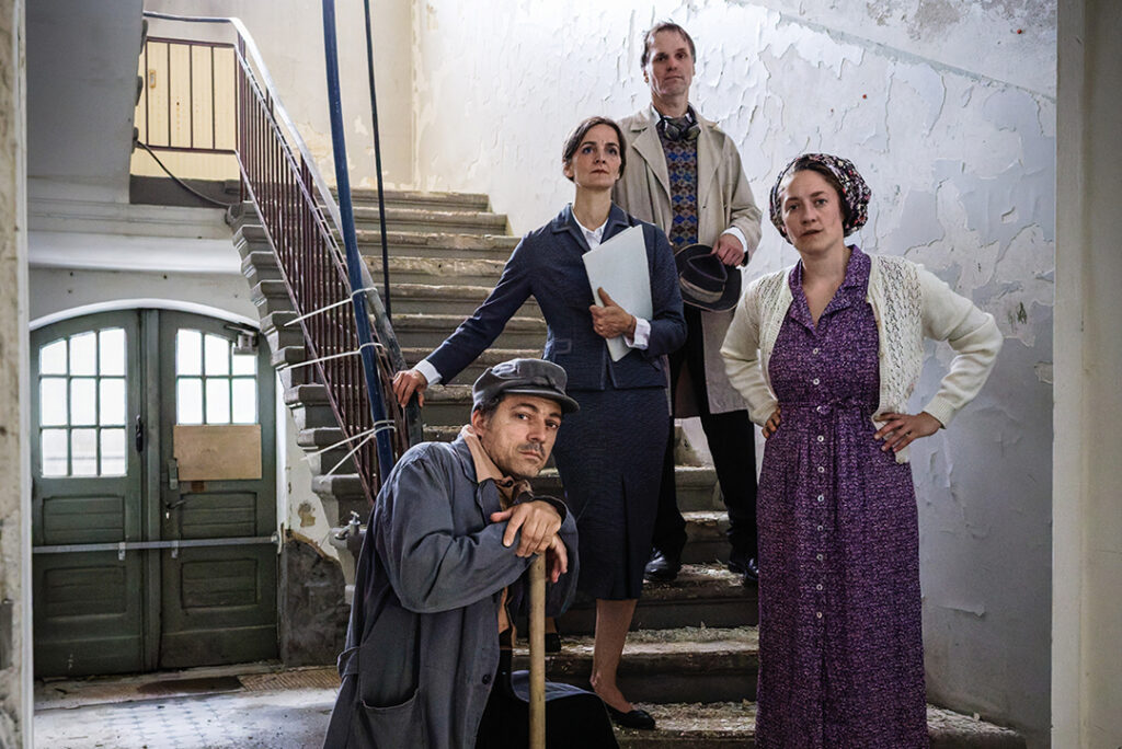 Vier Schauspieler in Kostümen posieren in einem heruntergekommenen Treppenhaus.