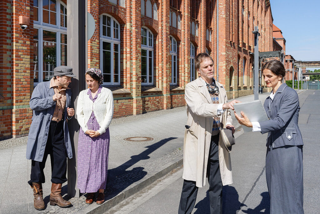 Schaupieler mit Kostümen aus den 1930er-Jahren posieren vor einem Backsteingebäude.
