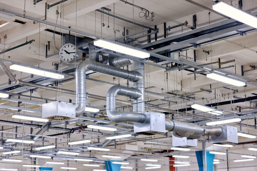 Blick an die Decke einer Fabrikhalle mit zahlreichen Lichtern, Kabeln, Verstrebungen und Rohren.