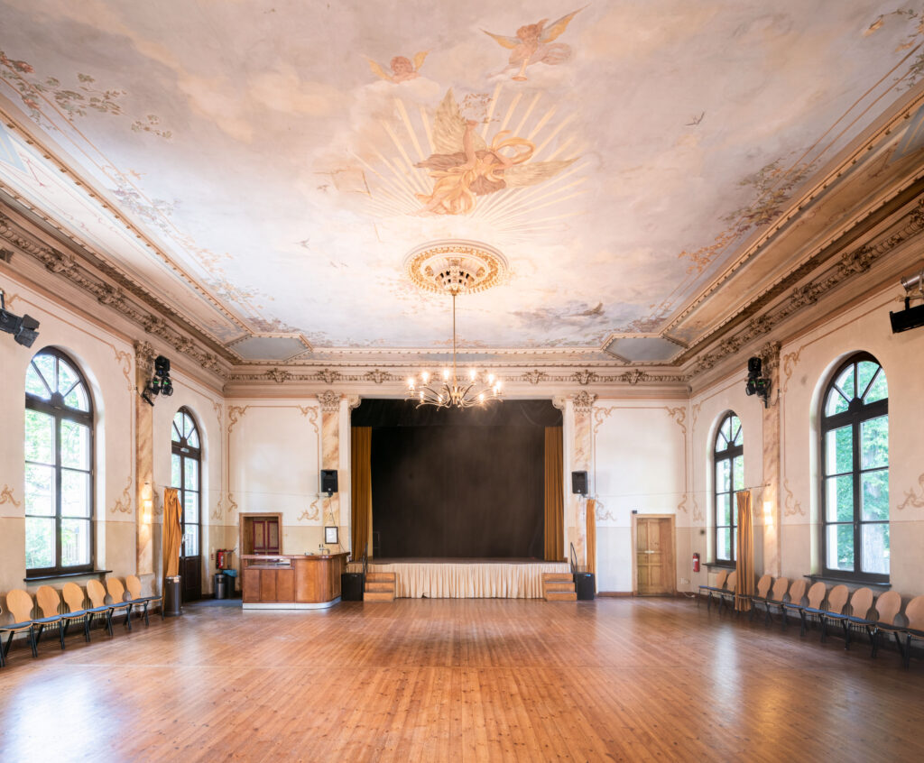 Saal mit leerer Tanzfläche. Am Rand stehen aufgereiht Stühle, die Bühne ist dunkel und leer. An der Decke hängt ein prunkvoller Leuchter und die Wände sind aufwendig verziert.