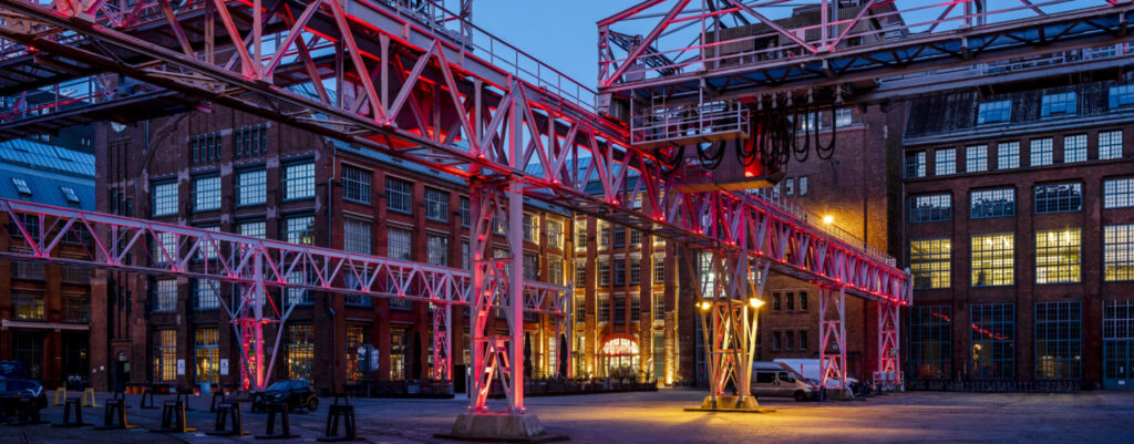 Krananlage vor historischem Fabrikgebäude am Abend, gelb und pink beleuchtet