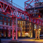 Krananlage vor historischem Fabrikgebäude am Abend, gelb und pink beleuchtet