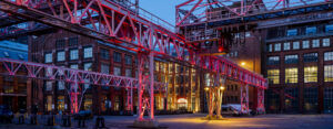 Krananlage vor historischem Fabrikgebäude am Abend, gelb und pink beleuchtet
