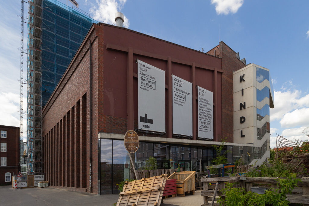 Außenansicht eines Backsteingebäudes vor blauem Himmel. Seitlich angebaut ist ein Treppenhaus aus Beton, auf dem in großen Lettern KINDL steht. Ein Teil des Gebäudes ist mit einem Baugerüst verkleidet.