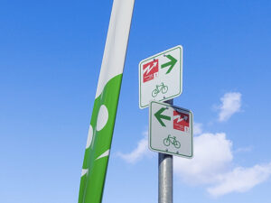 Roter Wegweiser der Radroute "Warmes Licht und kühles Bier", montiert an einem Fahrradwegweiser in Berlin. Im Hintergrund ist der Schriftzug des Deutschen Technikmuseums zu sehen.