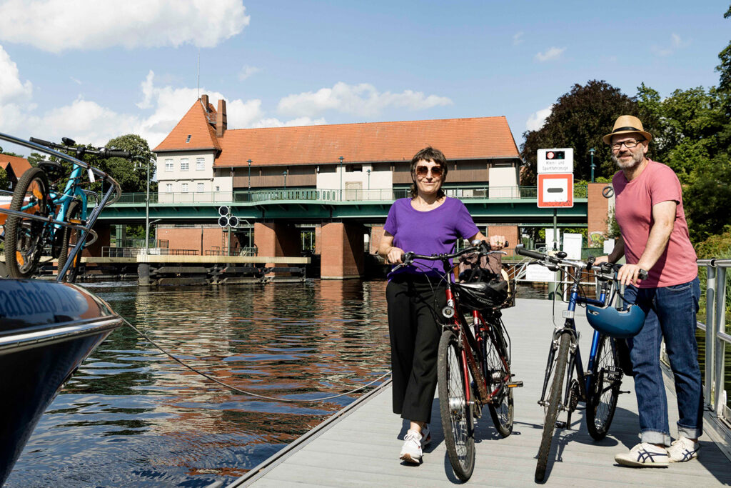 Zwei Radfahrende stehen auf einem Steg am Wasser. Im Hintergrund die Schleuse Kleinmachnow.