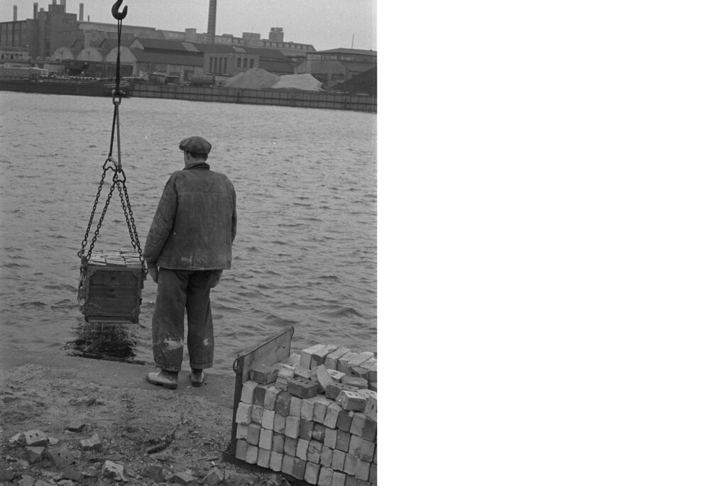 Schwarz-weiß Bild um 1955. Ein Arbeiter am Umschlagplatz in Schöneweide verlädt Ziegelsteine mit einem Kran an der Spree.