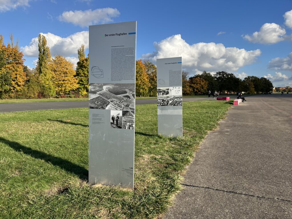 Das Foto zeigt zwei Infotafeln mit Texten und Fotos zum alten Flughafen Berlin, die entlang eines asphaltierten Wegs auf einer Grasfläche stehen.