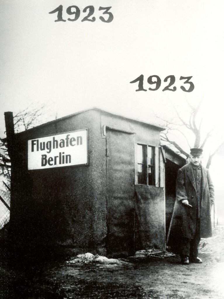 Historisches, schwarz-weißes Foto, das einen Mann vor einer kleinen Holzhütte zeigt. Auf der Holzhütte prangt ein Schild mit der Aufschrift Flughafen Berlin. Auf dem Foto ist zweimal die Jahreszahl 1923 zu sehen.