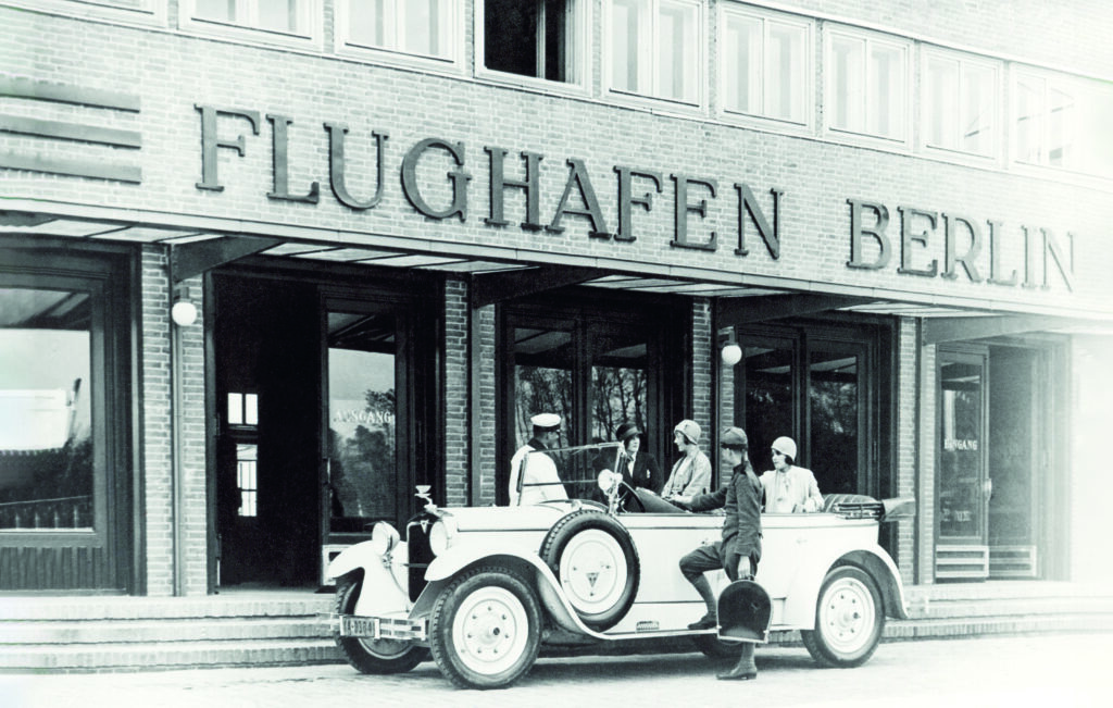 Ein schwarz-weißes, historisches Foto zeigt ein Auto mit offenem Verdeck, das vor einem Backsteingebäude mit dem Schriftzug FLUGHAFEN BERLIN hält. Einige Menschen sitzen im Auto bzw. stehen davor und dahinter.