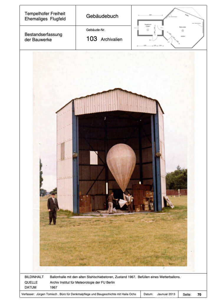 Das historische Foto zeigt einen weißen Wetterballon in der Ballonhalle. Am Ballon steht eine Person, eine weitere steht am linken Bildrand vor der Halle.