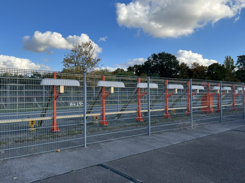 Hinter einem Metallzaun lassen sich rote Streben erkennen, an denen die elektrischen Leuchtfeuer befestigt sind.