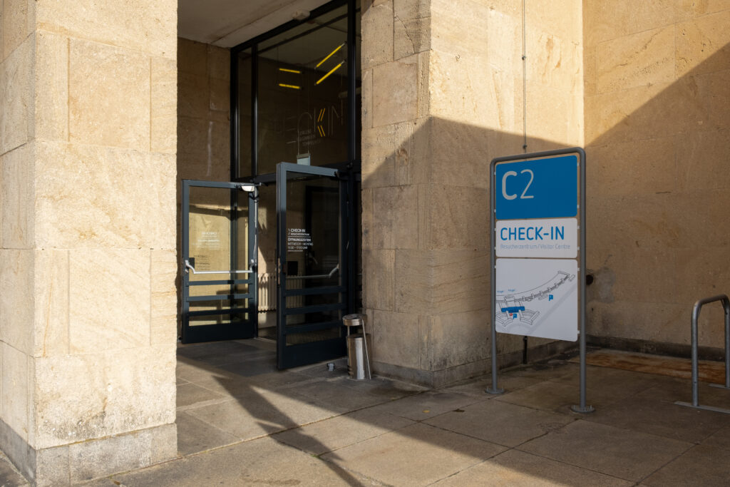 Das Foto zeigt den Eingang ins Besucherzentrum. Die Tür dazu steht offen, davor weist ein weiß-blaues Schild auf das Besucherzentrum hin. Die Wände des Gebäudes sind mit hellem Naturstein verkleidet.