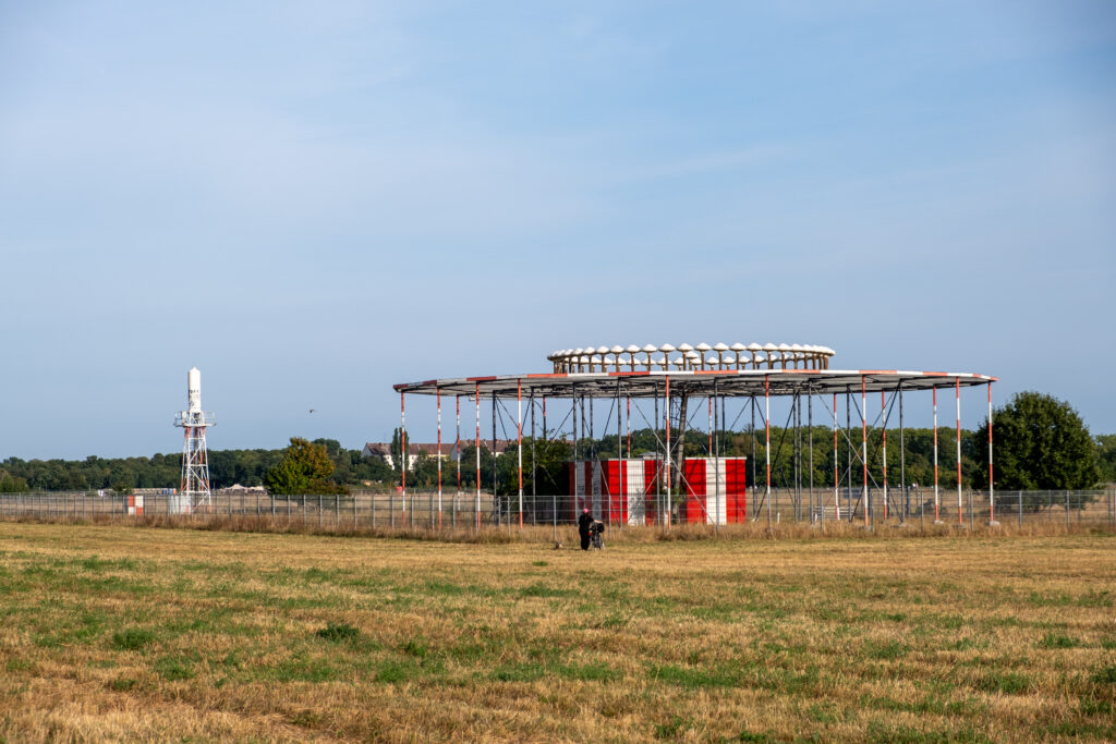 Das Foto zeigt das von einem Zaun umgebene Drehfunkfeuer (DVOR) auf der Rasenfläche des Tempelhofer Felds. Das Drehfunkfeuer besteht aus einer niedrigen runden Konstruktion aus rot-weißen, dünnen Metallstäben.