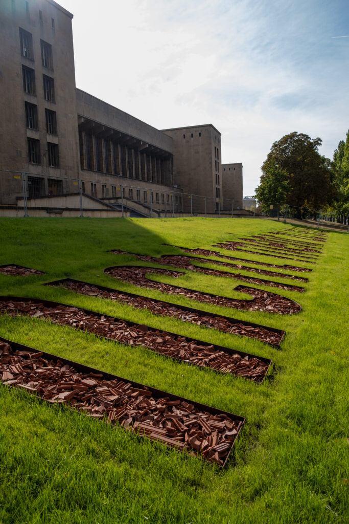 Foto der grünen Rasenfläche vor dem Flughafengebäude. Es sind große Buchstaben im Gras zu erkennen, die anscheinend mit losen Holzstücken befüllt sind. Die Buchstaben verjüngen sich perspektivisch zum Horizont hin, der komplette Schriftzug ist nicht lesbar.