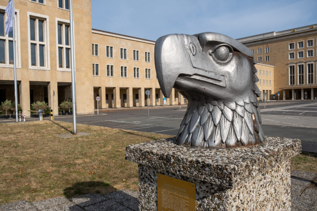 Der metallene Adlerkopf steht auf einem Sockel aus Waschbeton auf dem Vorplatz des Ehrenhofs des Flughafens. An dem Sockel befindet sich auch eine Texttafel.