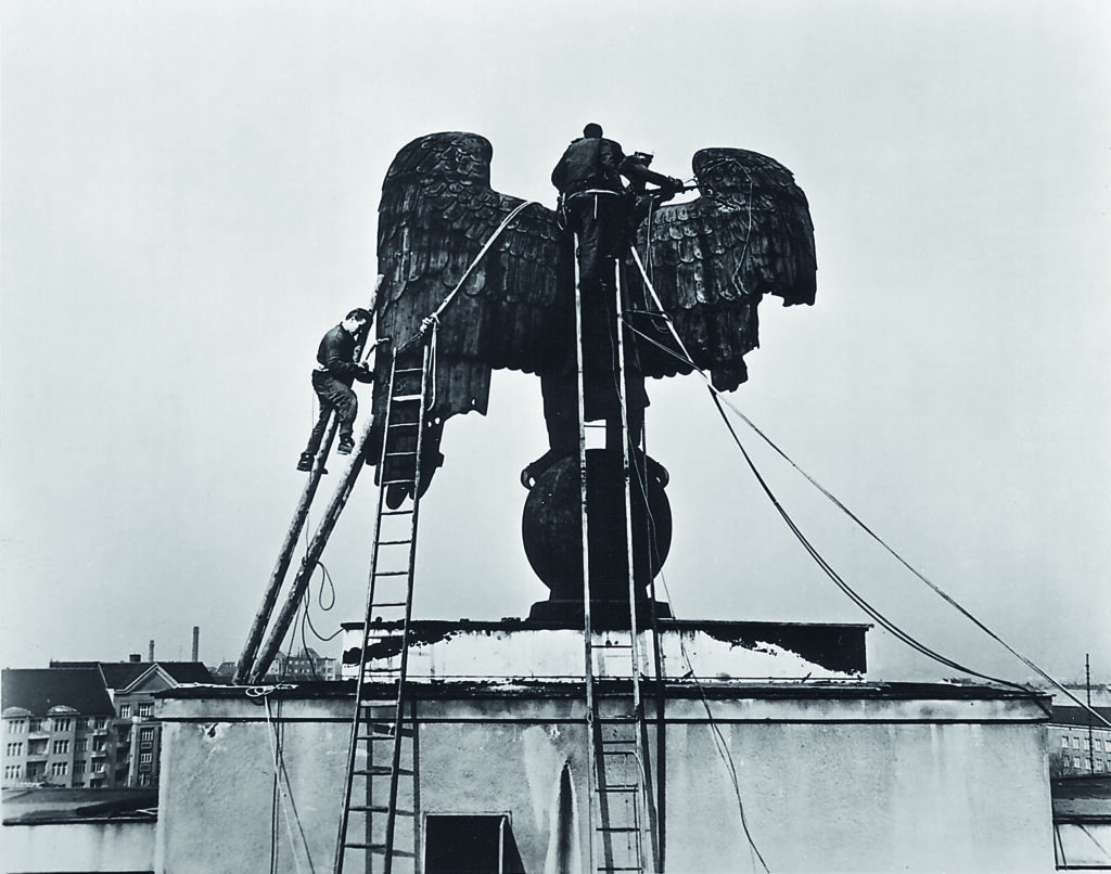 Ein historisches Foto in schwarz-weiß zeigt, wie die alte Adlerskulptur von zwei Personen auf Leitern demontiert wird.