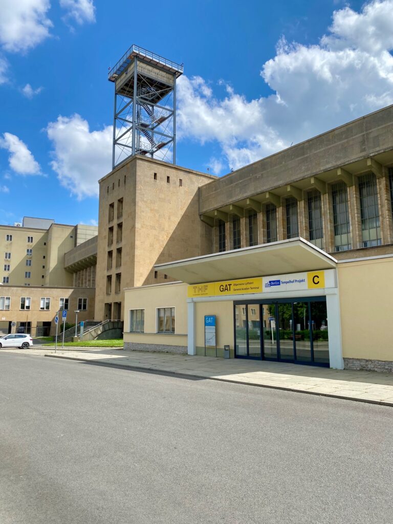 Das hochkant aufgenommene Foto zeigt den Eingang zum ehemaligen General Aviation Terminal mitsamt der Straße davor. Im Hintergrund sieht man blauen Himmel mit einigen weißen Wolken.