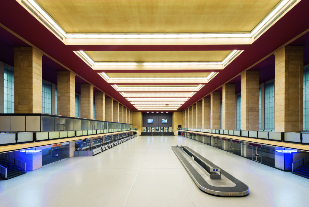 Weitwinklig aufgenommenes Foto der hohen Eingangshalle (Abfertigungshalle) im Flughafen Tempelhof. Markant sind die dicken rechteckigen Säulen, die die Decke stützen. In der Mitte des Raums sieht man ein Gepäckband.