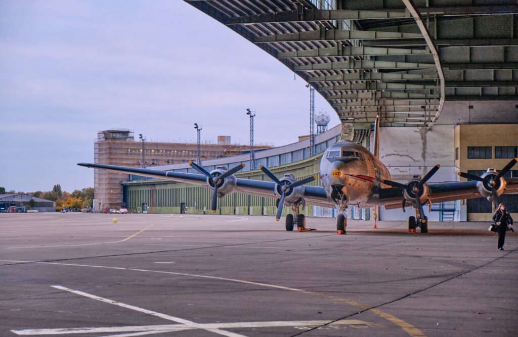 Eine silbern-glänzende Propellermaschine steht teilweise unter der Überdachung des Hangars, der eine elegante Kurve nach hinten links beschreibt. Im Hintergrund sieht man Teile des Rollfelds sowie den ehemaligen Fluglotsentower.