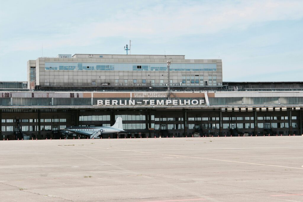 Das Foto zeigt die horizontale Ausdehnung des Flughafengebäudes mit dem überdachten Hangar davor. Vor dem Hangar steht ein Flugzeug. Im Vordergrund sieht man das Rollfeld.