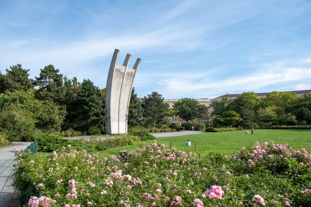 Das Foto zeigt das grau aufragende Luftbrückendenkmal inmitten einer blühenden, parkähnlichen Landschaft. Im Hintergrund sieht man den blauen Himmel, im Vordergrund Büsche mit rosafarbenen Blüten.