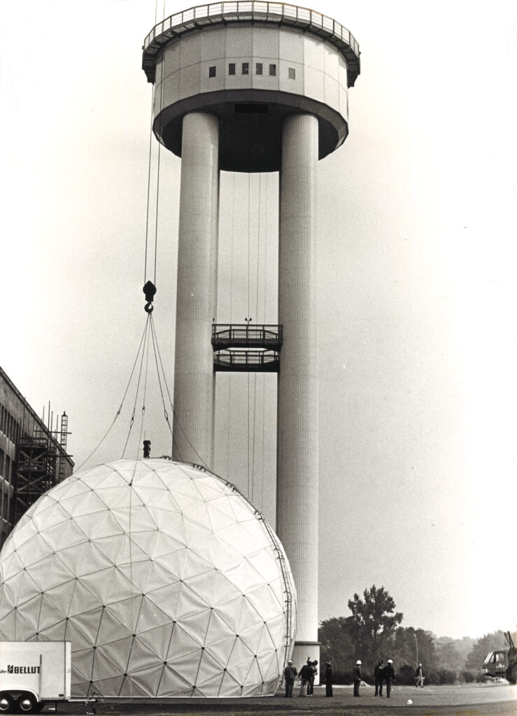 Historisches Foto in schwarz-weiß, das den Bau des Radarturms zeigt. Der obere, kuppelförmige weiße Teil des Turms ist noch am Boden und wird scheinbar in wenigen Augenblicken von einem Kran hochgezogen.