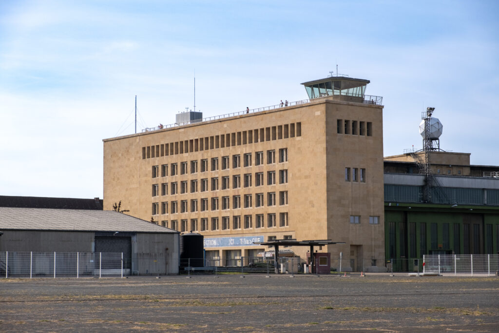 Außenansicht des ehemaligen Fluglotsentowers. Das längliche, rechteckige Gebäude besteht aus hellbraunem Stein und überragt die beiden anderen Gebäude links und rechts von ihm.