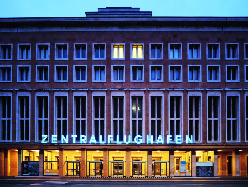 Frontales Foto, das das Flughafengebäude mit dem beleuchteten Schriftzug ZENTRALFLUGHAFEN zeigt. Der Eingang sowie zwei Fenster im obersten Stock sind erleuchtet.