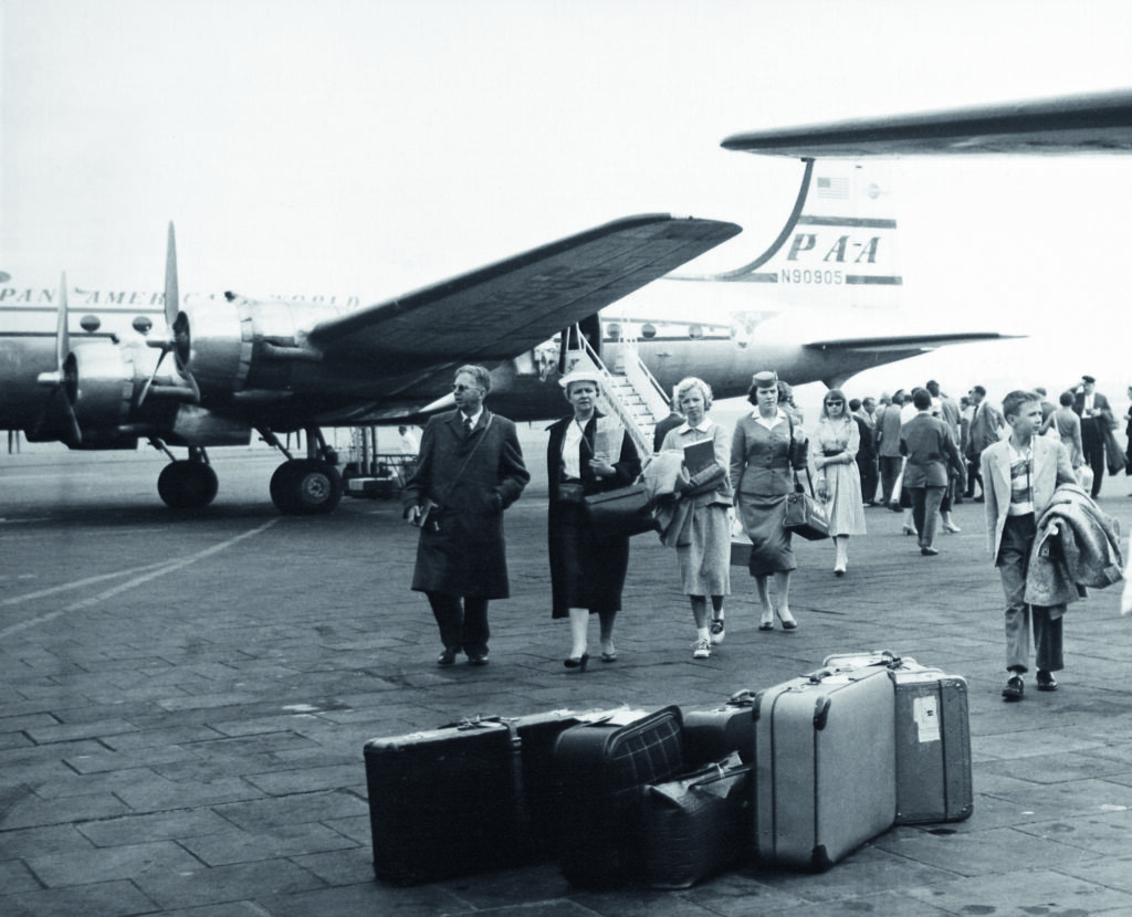 Historisches Foto in schwarz-weiß, das Menschen nach dem Aussteigen aus einer Propellermaschine zeigt. Im Vordergrund stehen einige Reisekoffer.