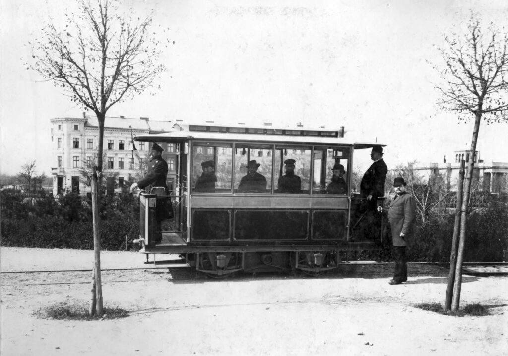 Historische schwarz-weiß-Aufnahme einer Straßenbahn. Im kurzen Waggon sitzen mehrere Männer mit Hut. Auch auf den beiden Außenplattformen an den Enden stehen Personen.