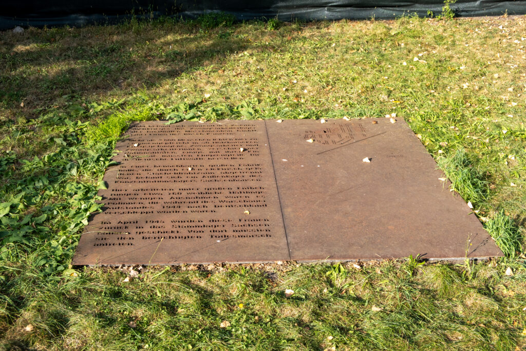 Foto einer Gedenkplatte im Gras. Auf dem linken Teil der Platte ist Text zu sehen, der aber aufgrund der Entfernung nicht lesbar ist.