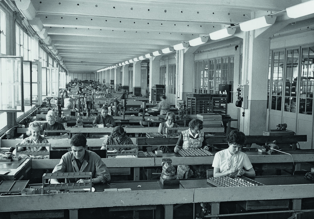Historisches schwarz-weißes Foto, das eine große Halle zeigt, in der viele Arbeiterinnen und Arbeiter Optikteile herstellen.