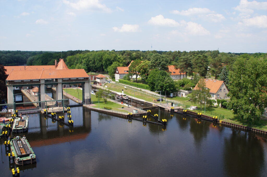 Luftaufnahme der Schleuse Kleinmachnow. Der Himmel ist blau und die Ufer sind bewaldet mit einigen vereinzelten Häusern dazwischen.