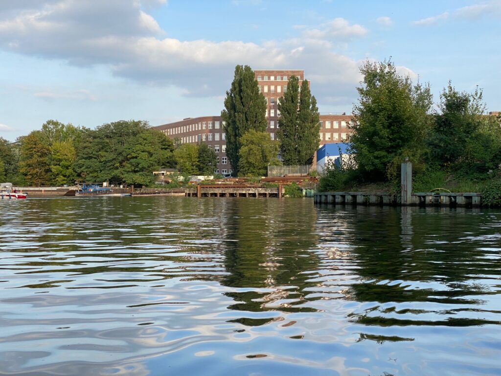 Ansicht des Gebäudekomplexes des Radio Loewe vom Wasser des Teltowkanals aus. Einige Bäume verdecken das Gebäude.