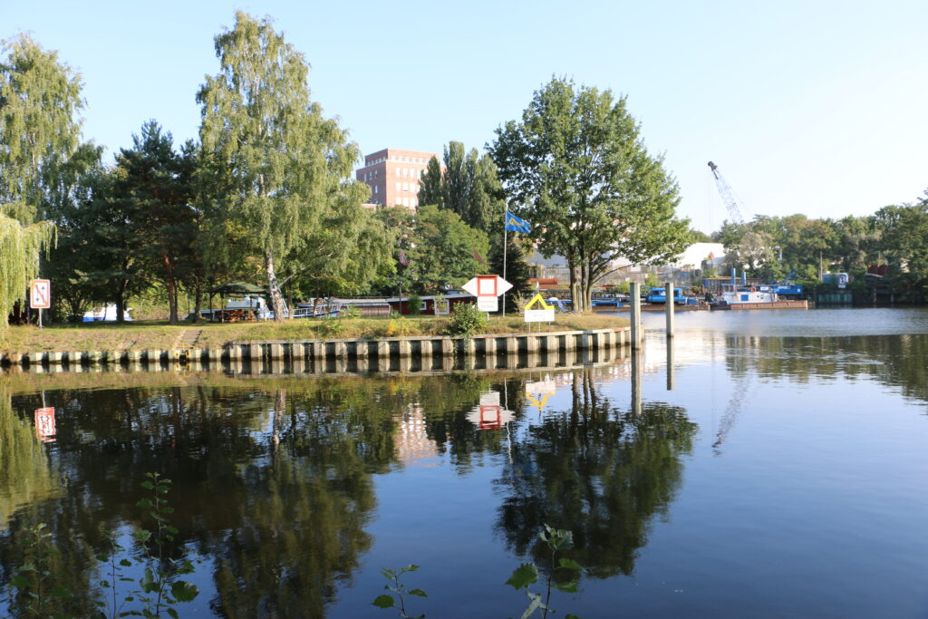 Der Hafen Steglitz vom Wasser aus gesehen. Grüne Bäume und blauer Himmel spiegeln sich im Wasser des Teltowkanals.