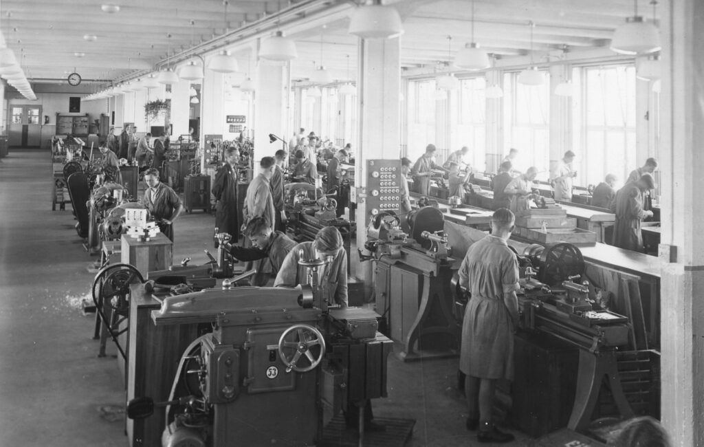Historische, schwarz-weiße Aufnahme von Arbeiterinnen und Arbeitern im damaligen Telefunken-Werk. In einer großen, lichtdurchfluteten Halle sieht man viele Menschen an Maschinen arbeiten.