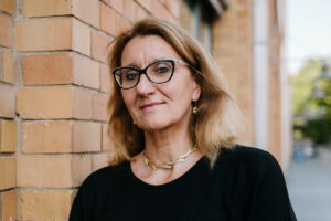 Teamfoto von Susanne Kähler. Sie lehnt an einer Backsteinwand und blickt in die Kamera.